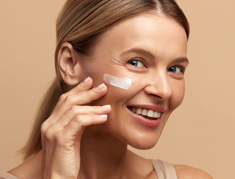 Lächelnde Frau mittleren Alters trägt eine weiße Creme auf ihre Wange auf.